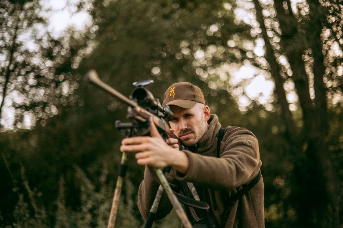 A hunter in an outdoor setting aiming a rifle while wearing a cap and jacket, with trees blurred in the background.