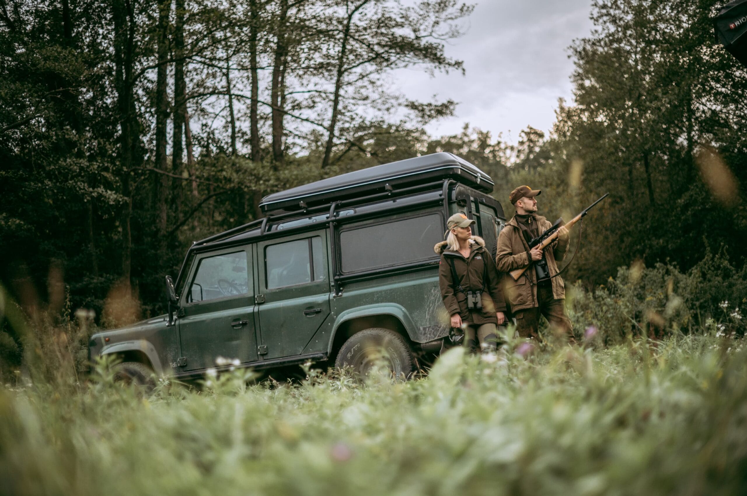 Two hunters stand beside a green Land Rover, showcasing a moment of camaraderie in a Hungarian forest.